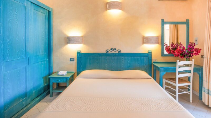 Camera da letto con mobili turchesi, letto matrimoniale beige, specchio e vaso con fiori rosa su scrivania.