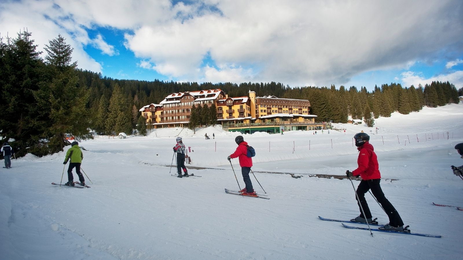 Group of skiers descending a snowy slope with chalets and trees in the background, cloudy sky.