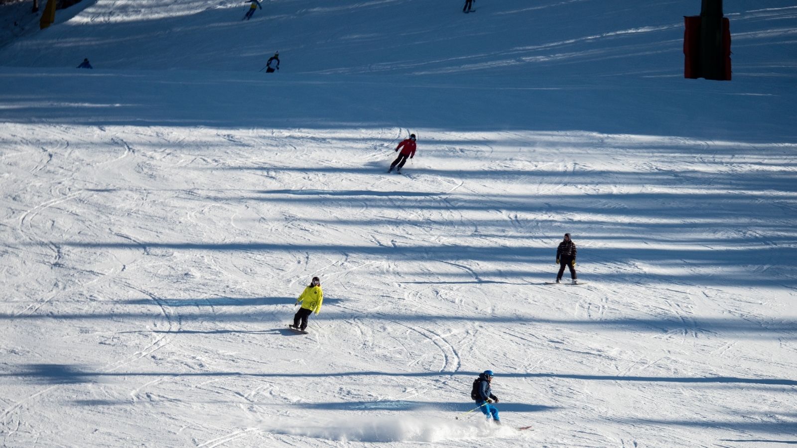 Skiers descend a snow slope with long shadows and ski tracks under a clear sky.