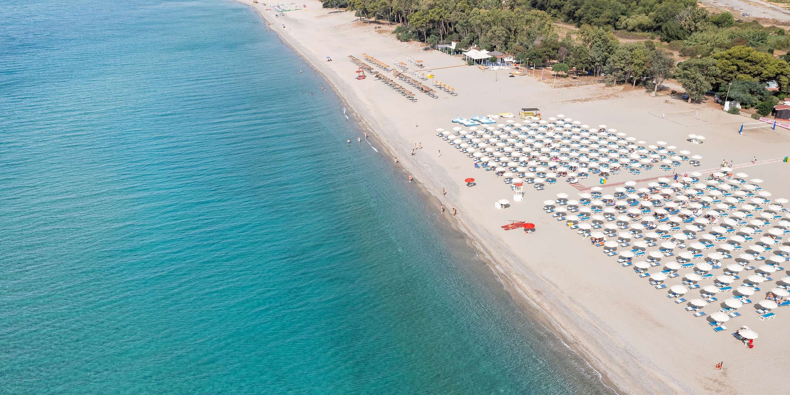 Plaża z uporządkowanymi parasolami i leżakami, turkusowe morze i ludzie na brzegu w słoneczny dzień.