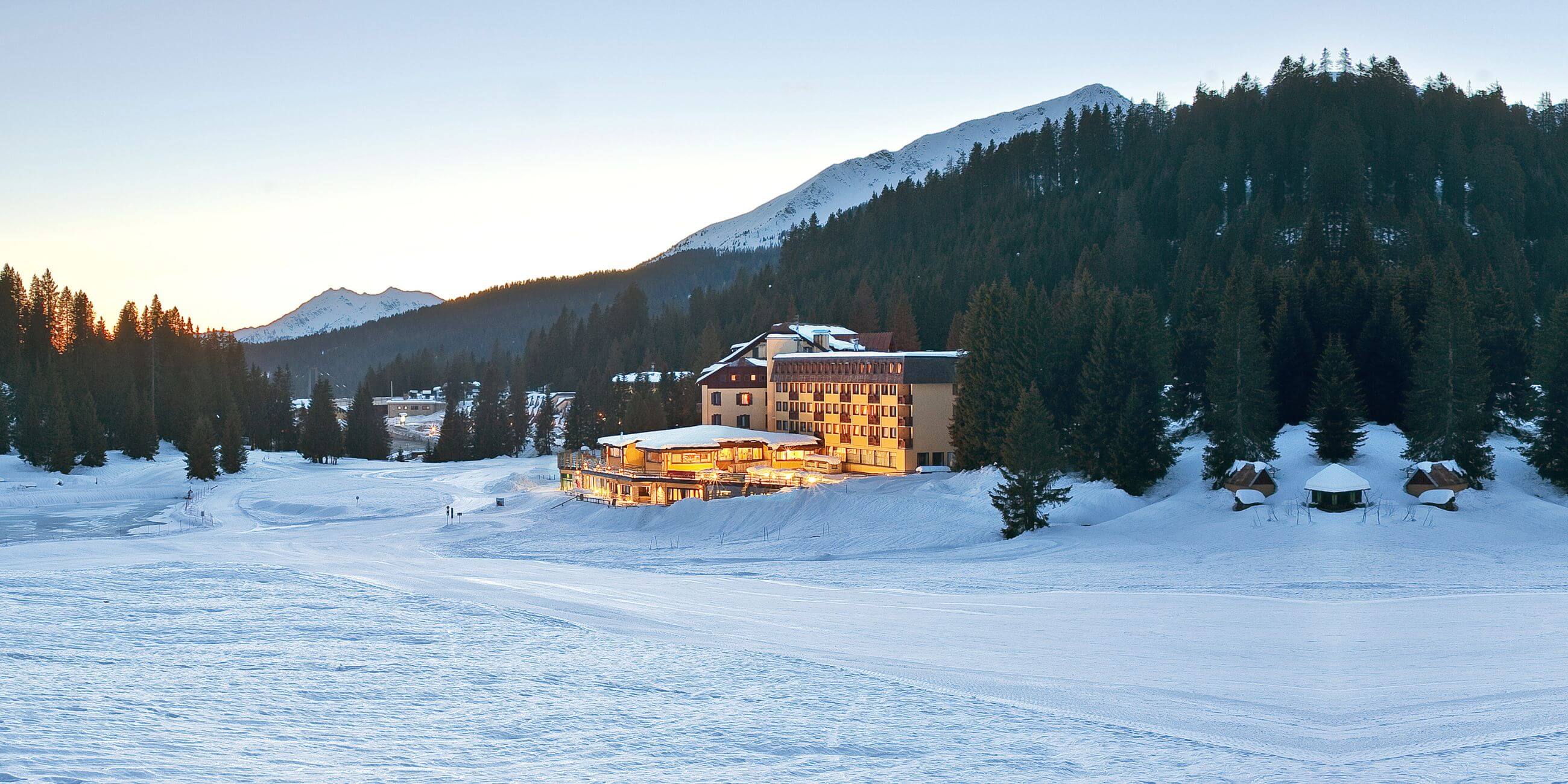Illuminated winter hotel at the foot of a snowy mountain, surrounded by trees and an alpine landscape at sunset.