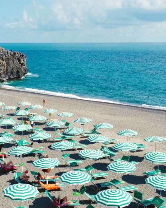 Beach with green and white umbrellas arranged on gray sand near a cliff and blue sea.