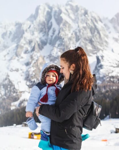 A mother is holding a winter baby in her arms, with snow-covered mountains in the background. Both are smiling happily.
