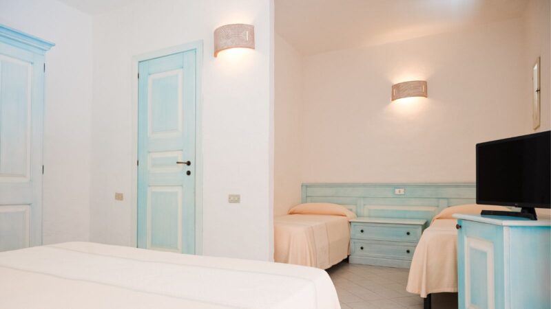 Camera con tre letti singoli, mobili e porta in legno azzurro e pareti bianche illuminate da applique.