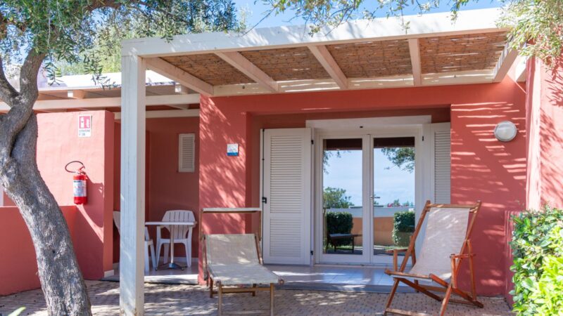 Veranda con due sedie a sdraio e tavolo con sedie bianche davanti a casa rosa con tettoia e albero.