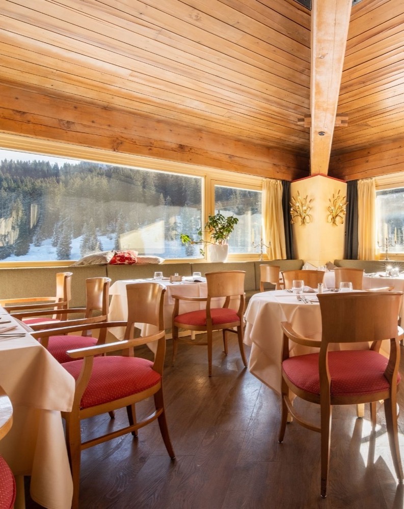 Interno di ristorante con grandi vetrate che mostrano un paesaggio innevato, tavoli con tovaglie e sedie con cuscini rossi.