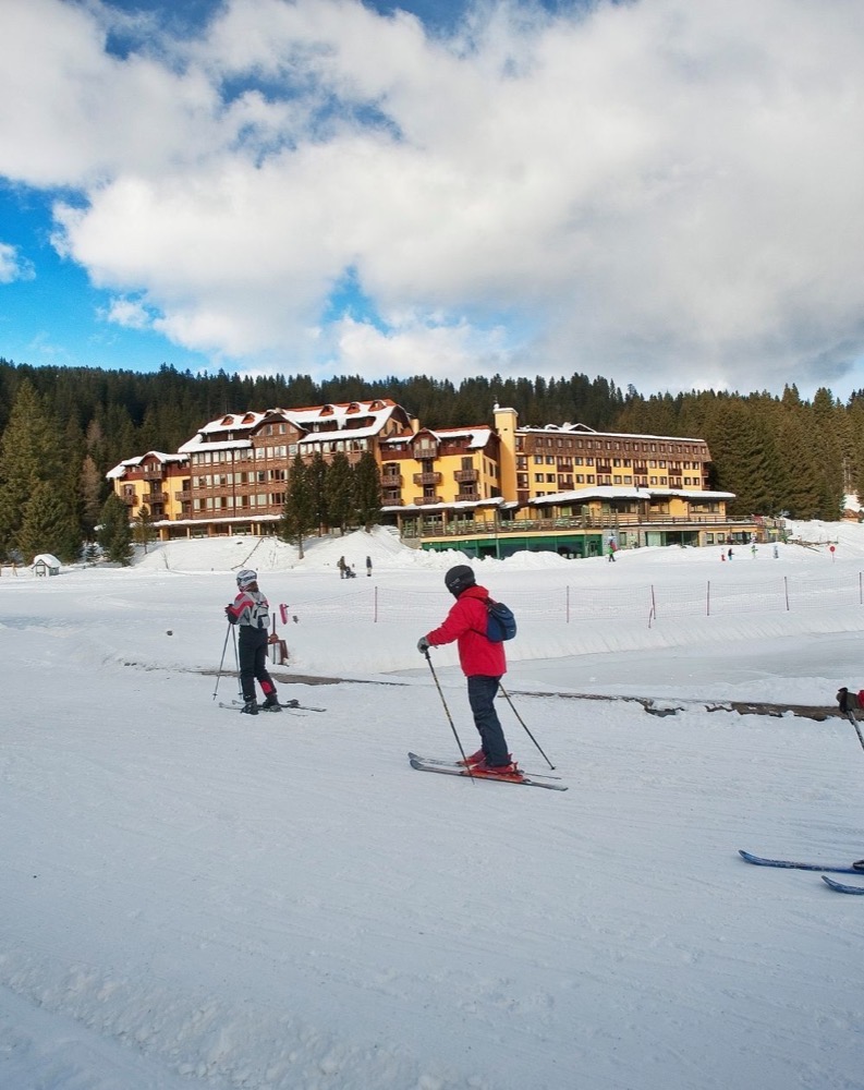Sciare sulla neve con un grande hotel di montagna sullo sfondo e cielo parzialmente nuvoloso.