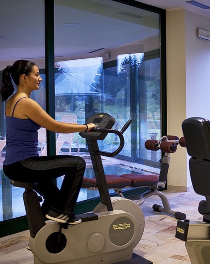 Donna che si esercita su una cyclette in palestra con grandi finestre che mostrano un paesaggio esterno con alberi.