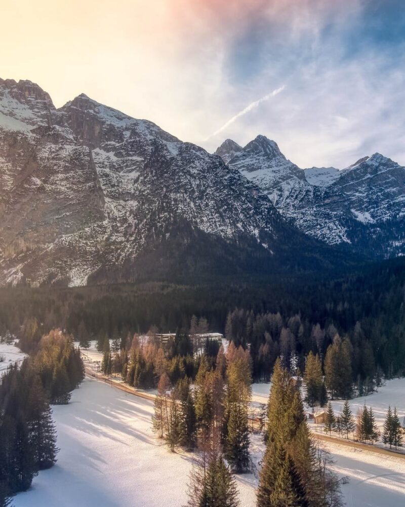 Paesaggio montano con alberi innevati, strada illuminata dal sole al tramonto e cime innevate sullo sfondo.