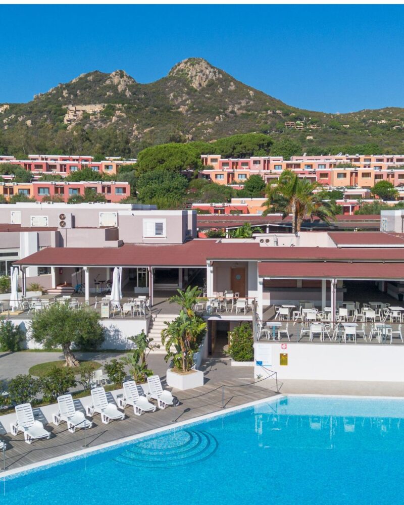 Vista panoramica di un resort con piscina, sdraio e colline sullo sfondo, in un ambiente soleggiato e accogliente.