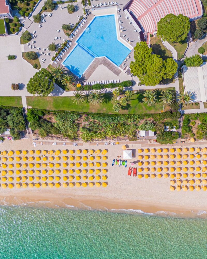 Vista aerea di una spiaggia con ombrelloni gialli, sabbia dorata e una piscina blu circondata da piante e lettini.