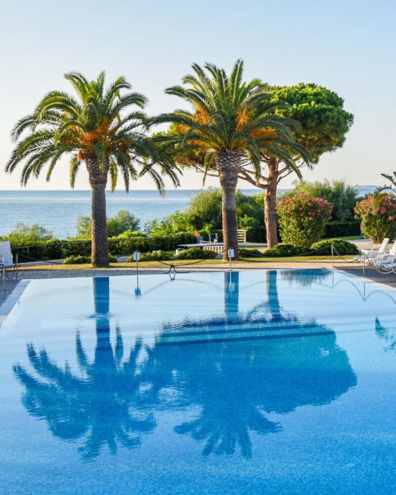 Piscina illuminata con palme e riflessi sull'acqua, vista sul mare e vegetazione fiorita intorno.