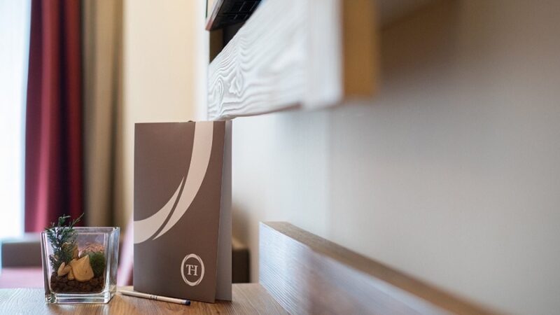 Dettaglio di un tavolo in legno con una broshure TH, una penna e una piccola decorazione con piante in vaso di vetro.