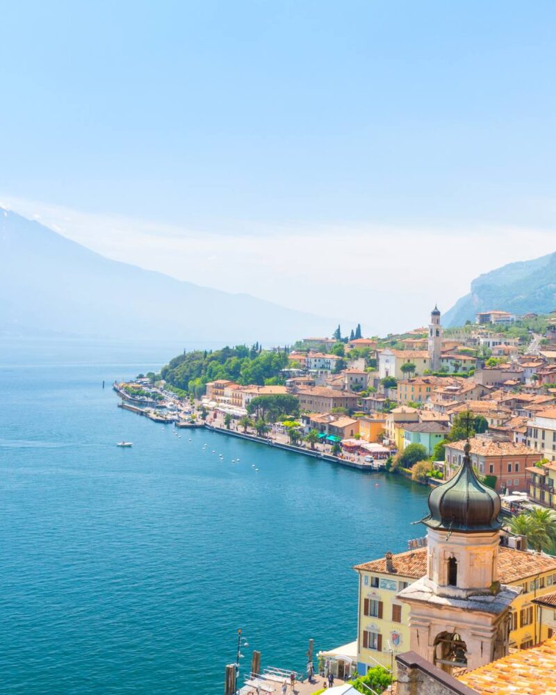 Vista panoramica del lago e borgo con tetti arancioni, campanili e montagne sullo sfondo, cielo azzurro terso.