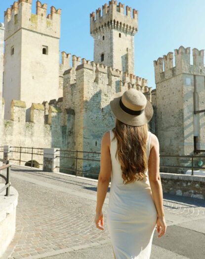 Donna con abito bianco e cappello estivo che guarda il castello medievale in una giornata di sole.