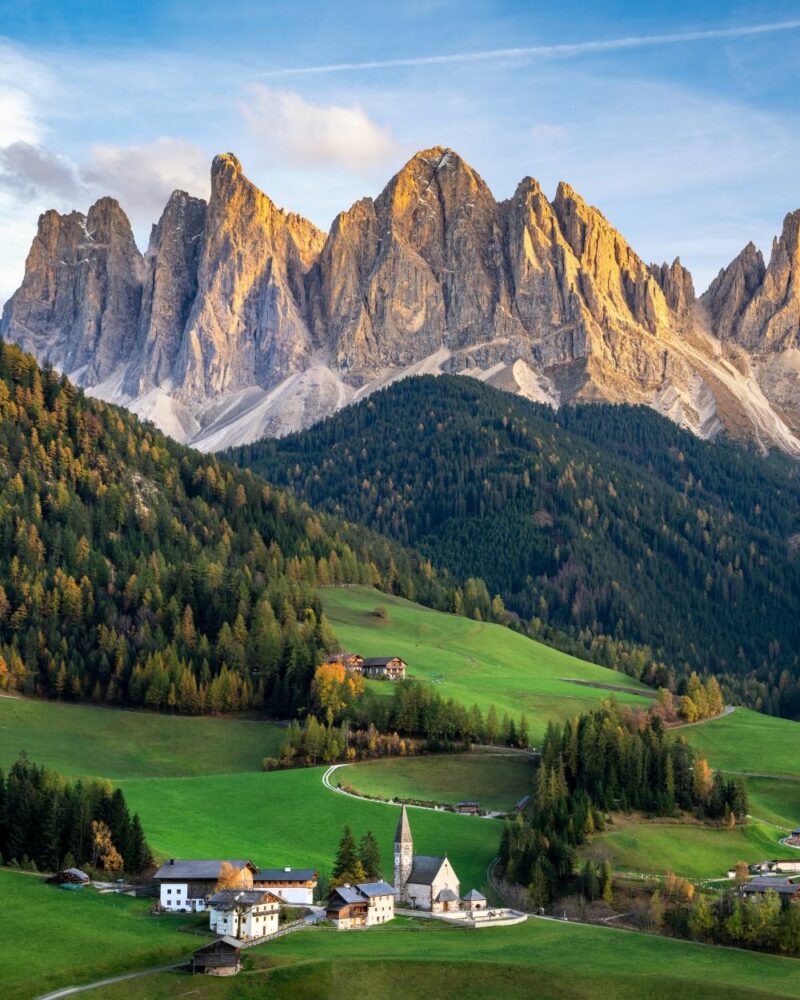 Paesino alpino con chiesa e case tra prati verdi, boschi e imponenti montagne rocciose illuminate dal sole.