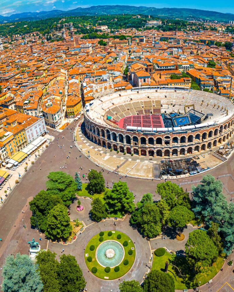 Veduta aerea dell'Arena di Verona e della città con tetti rossi e area verde con fontana in primo piano.