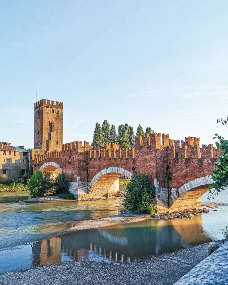 Ponte Scaligero rosso in mattoni con torri merlate sul fiume, riflesso nell'acqua e cielo sereno al tramonto.