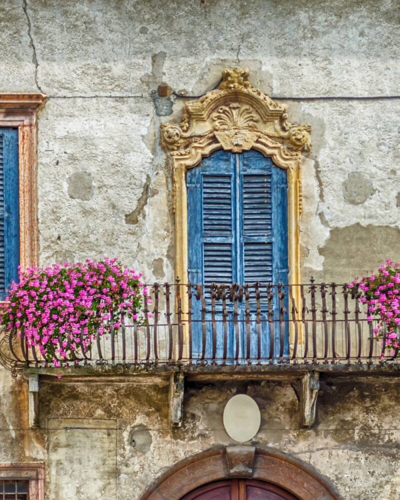 Balcone antico con infissi blu, fioriere con fiori rosa e muri in pietra sbiaditi.