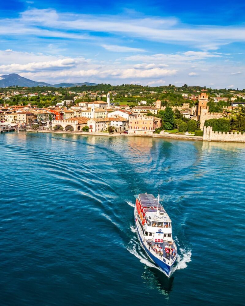 Battello turistico che naviga sul lago di fronte a un borgo storico con castello e colline verdi sullo sfondo.