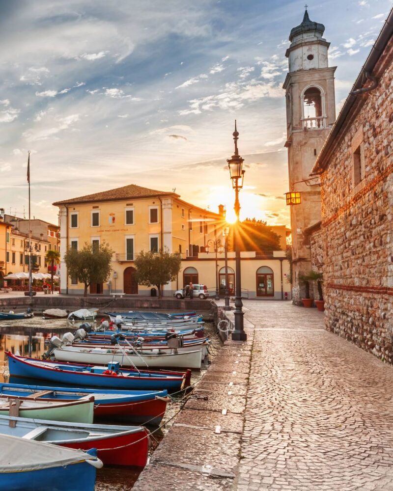 Barche colorate ormeggiate lungo il porto al tramonto con edificio storico e campanile sullo sfondo.