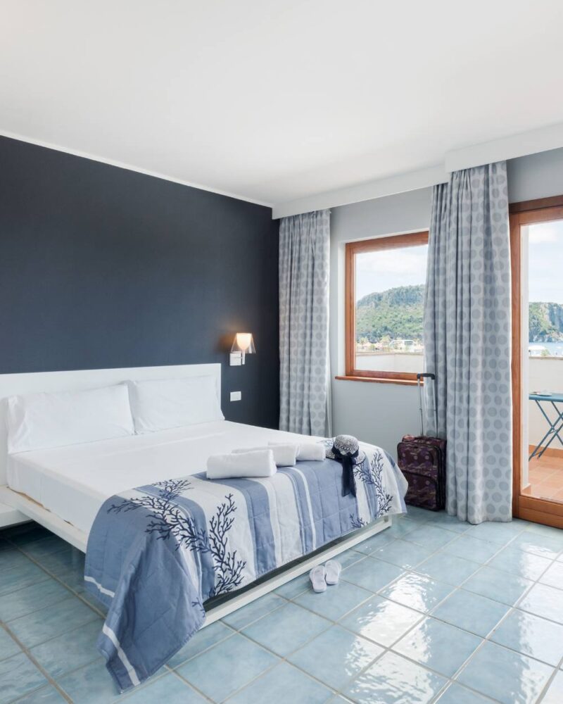 Camera da letto luminosa con letto matrimoniale, lenzuola bianche, copriletto blu, terrazza e vista panoramica sulle montagne.