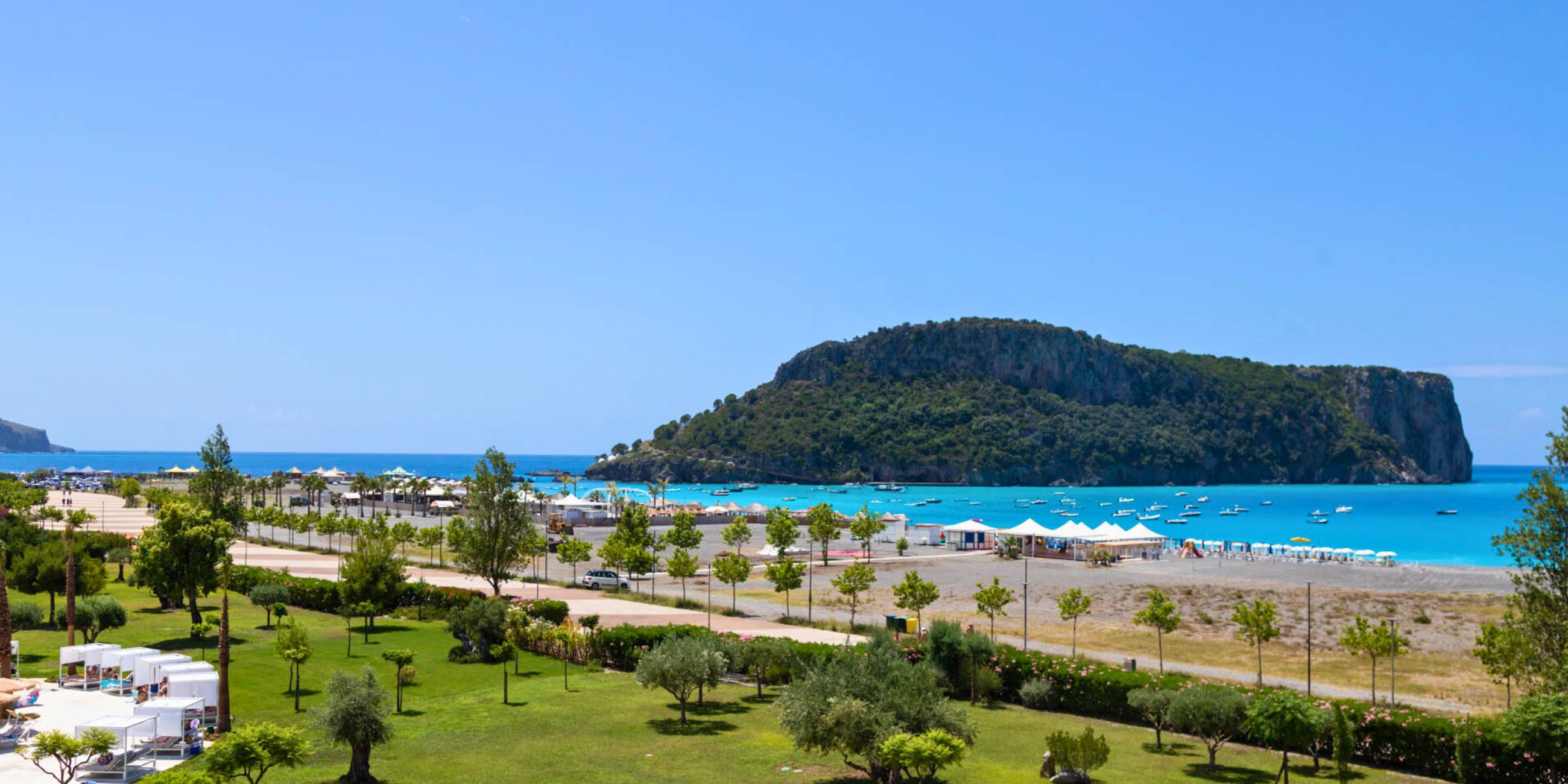 Spiaggia con ombrelloni bianchi, mare turchese e isola verde sullo sfondo sotto cielo azzurro sereno.