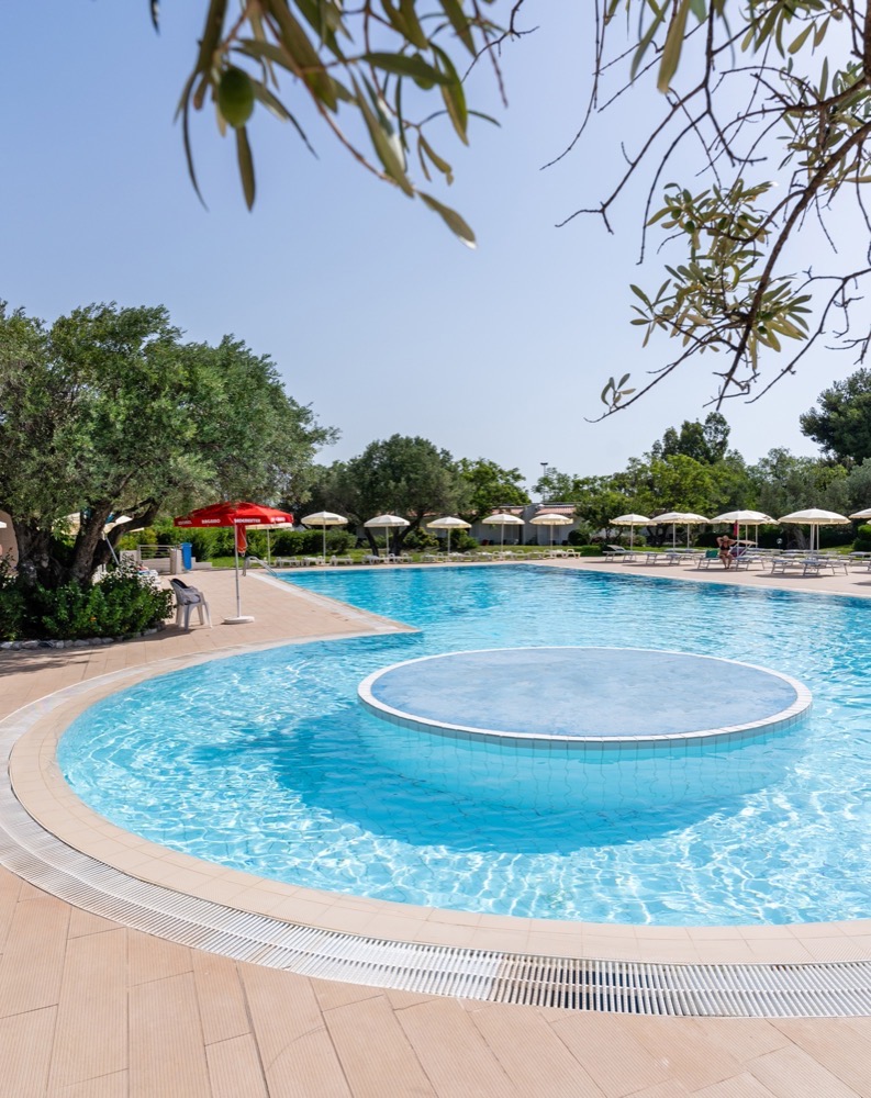 Piscina all'aperto con ombrelloni bianchi e alberi verdi sotto cielo sereno.