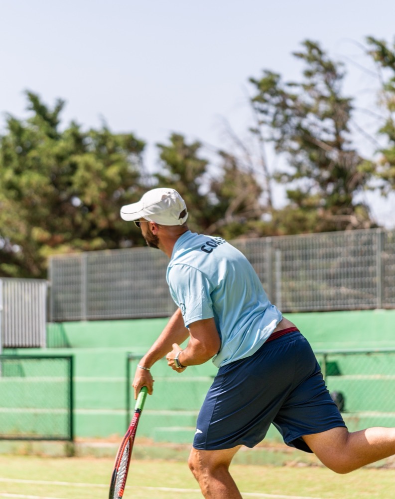Uomo con cappellino bianco che gioca a tennis su campo all'aperto con rete e alberi sullo sfondo.