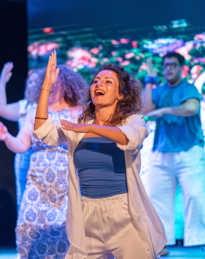 Donna sorridente che balla sul palco con altre persone, in abbigliamento casual chiaro sotto luci colorate.