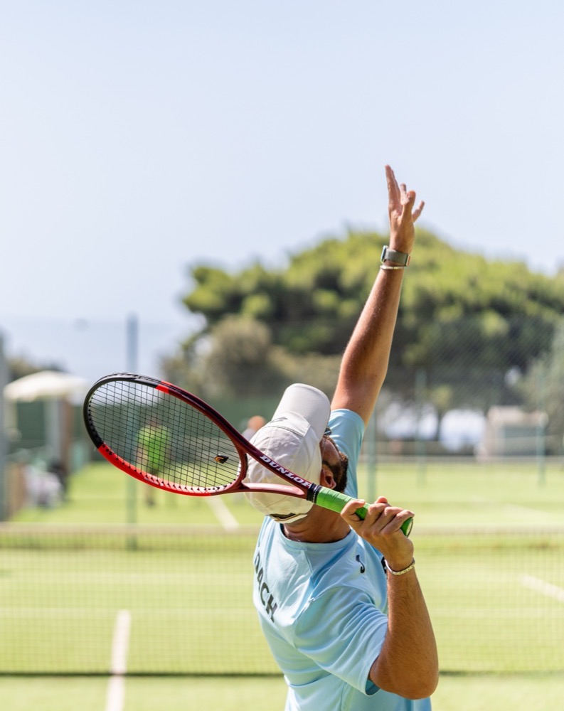 Uomo con cappello bianco e maglietta azzurra serve una palla su un campo da tennis all'aperto soleggiato.