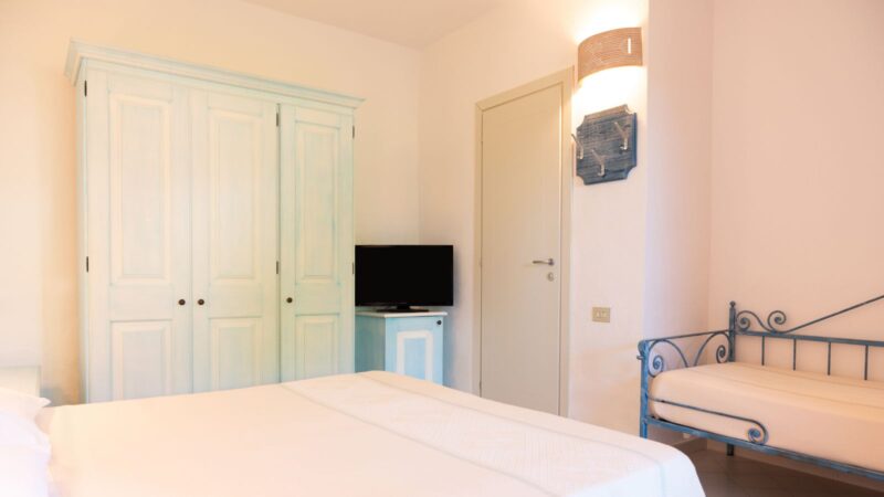 Camera luminosa con armadio bianco, letto matrimoniale con lenzuola bianche e divanetto in ferro battuto blu.