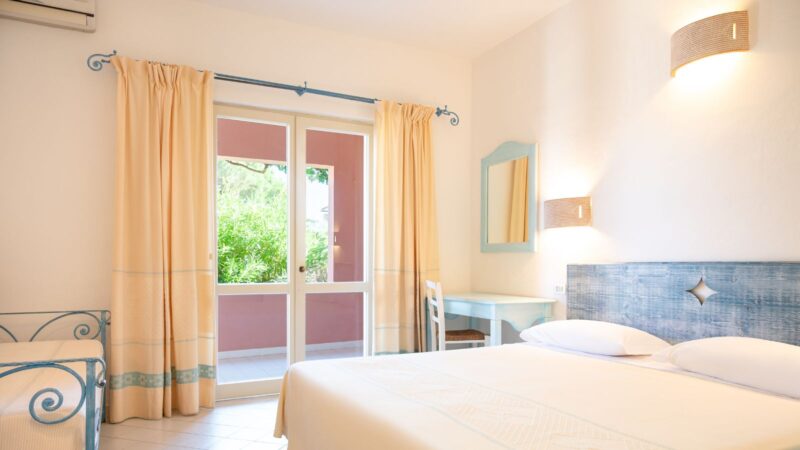 Camera luminosa con letto matrimoniale, scrivania azzurra, tende beige e porta finestra che dà su un giardino verde.