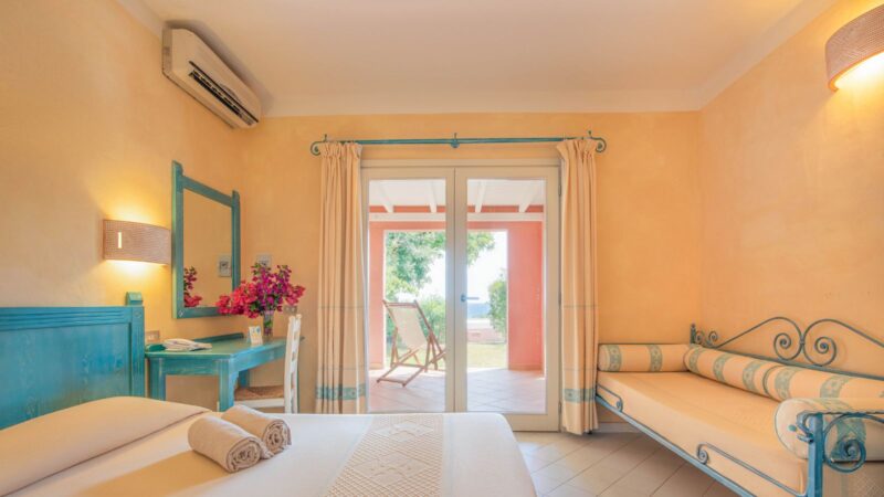 Camera d'hotel luminosa con letto matrimoniale, scrivania blu, divano beige e porta finestra con uscita su veranda.