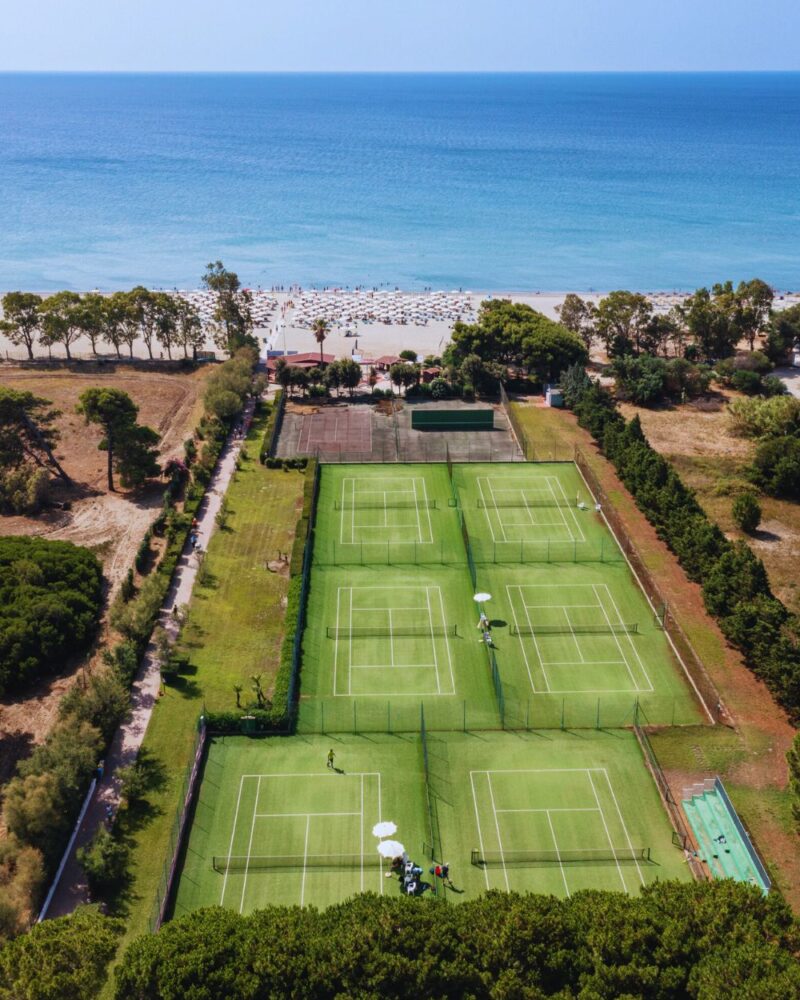 Campo da tennis circondato da vegetazione, con il mare e la spiaggia sullo sfondo. Sole e ombre dei parasole presenti.