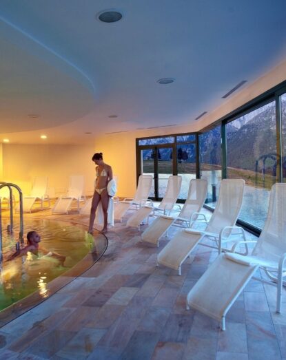 Exterior of an indoor pool with empty lounge chairs and a woman taking care of a child in the water.
