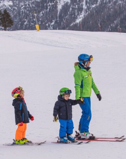 Children and ski instructor lined up on the snow, wearing helmets and winter clothing.