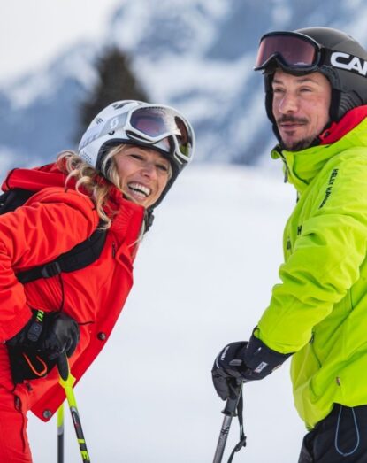 Two smiling skiers on the snow, wearing helmets and ski clothing.