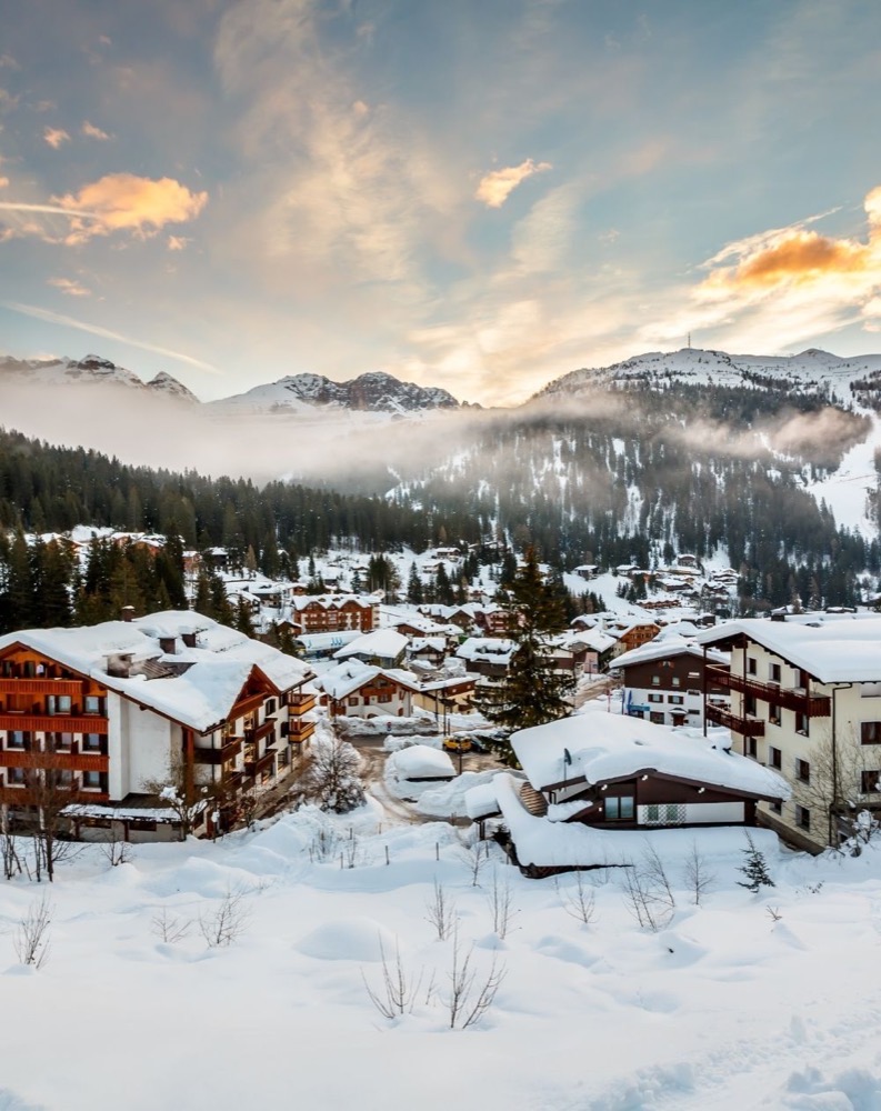 Paesaggio montano invernale con case e alberi coperti di neve, al tramonto con cielo nuvoloso.