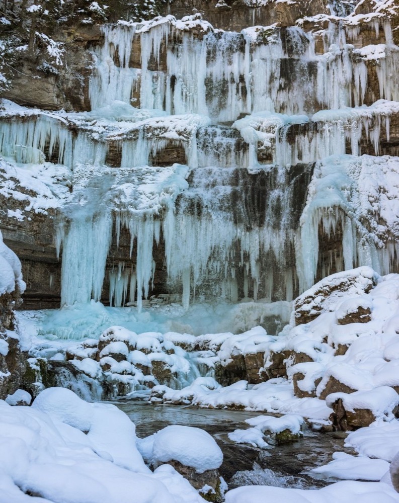 Cascata ghiacciata con neve e rocce, circondata da natura invernale.