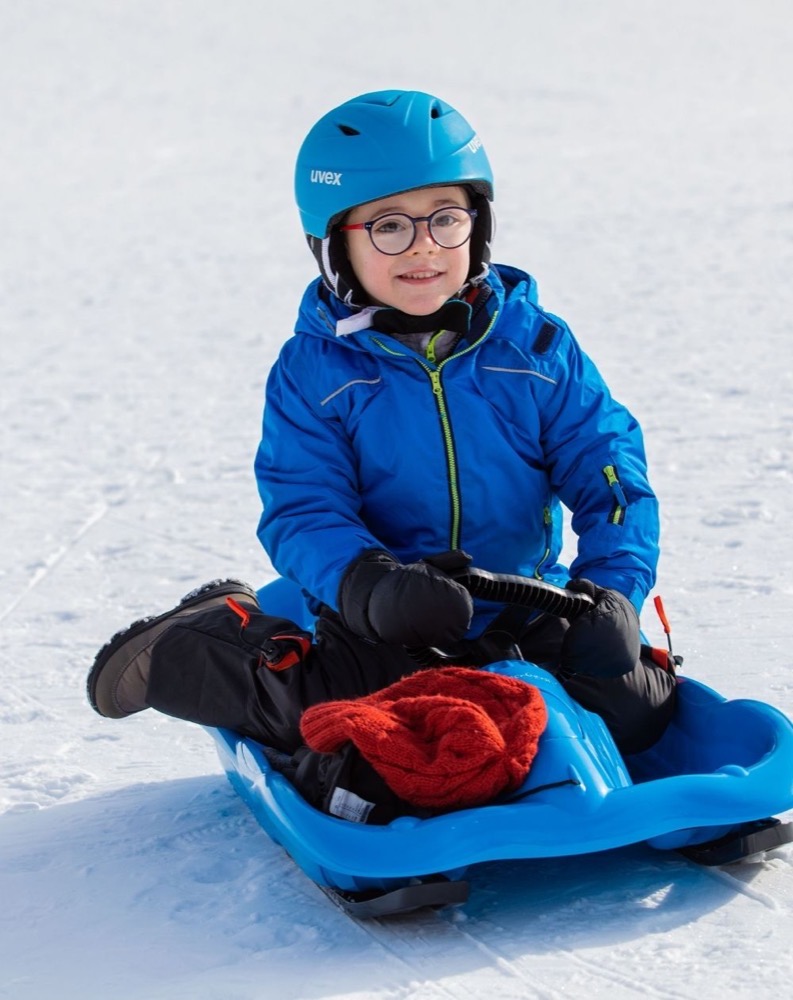 Ragazzo con casco e occhiali, seduto su una trottola sulla neve, indossa giacca blu e tiene una coperta rossa.