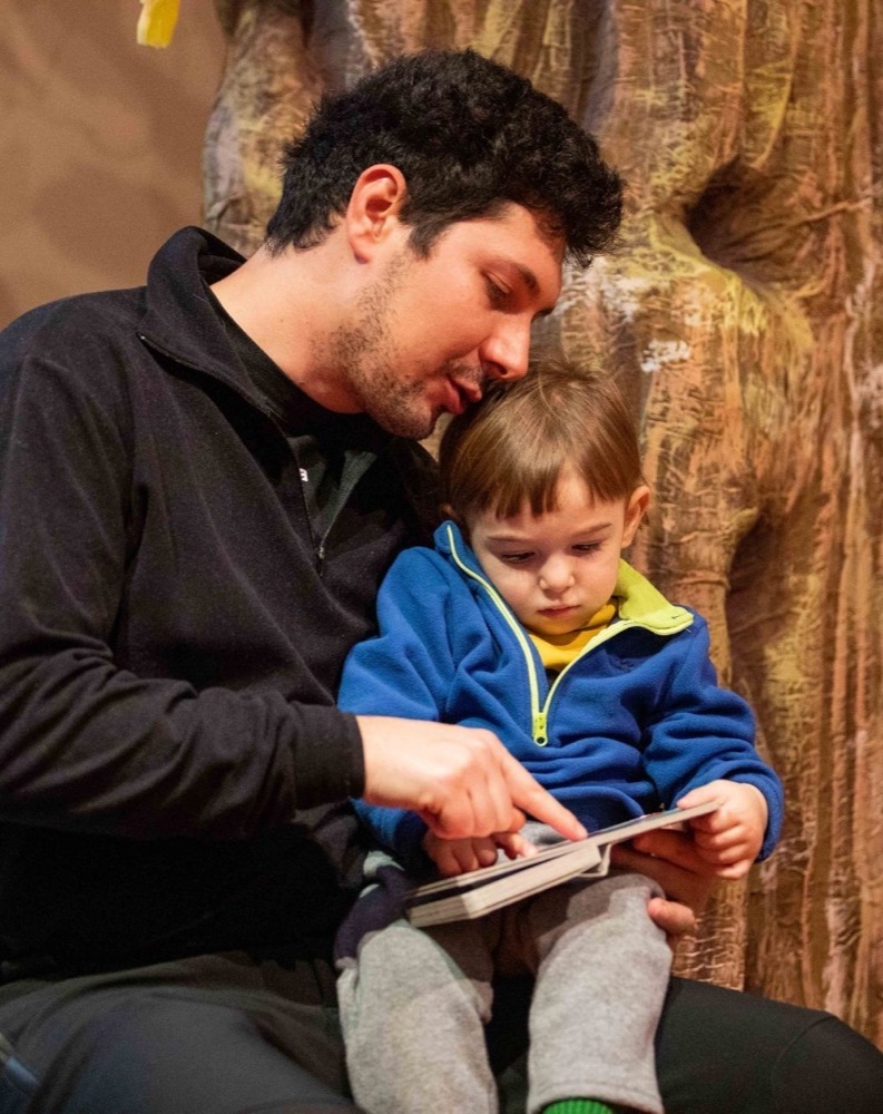 Uomo e bambino legati, che leggono insieme un libro intorno a un ambiente naturale.