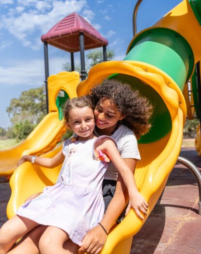 Dwie uśmiechnięte dziewczynki zjeżdżają razem ze żółtej zjeżdżalni na słonecznym placu zabaw.