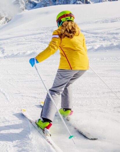 Girl with a green helmet and yellow jacket skiing on the snow with mountains in the background.