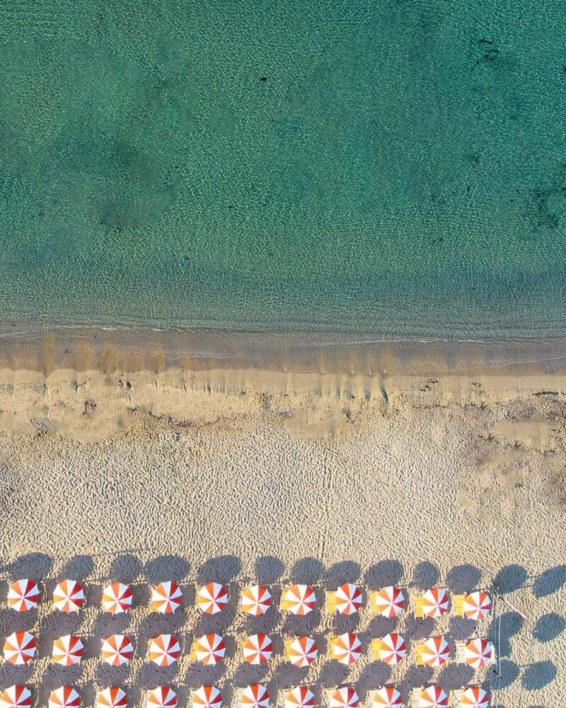 Spiaggia con lettini e ombrelloni rossi e bianchi, acque cristalline e sabbia chiara che incontra il mare.