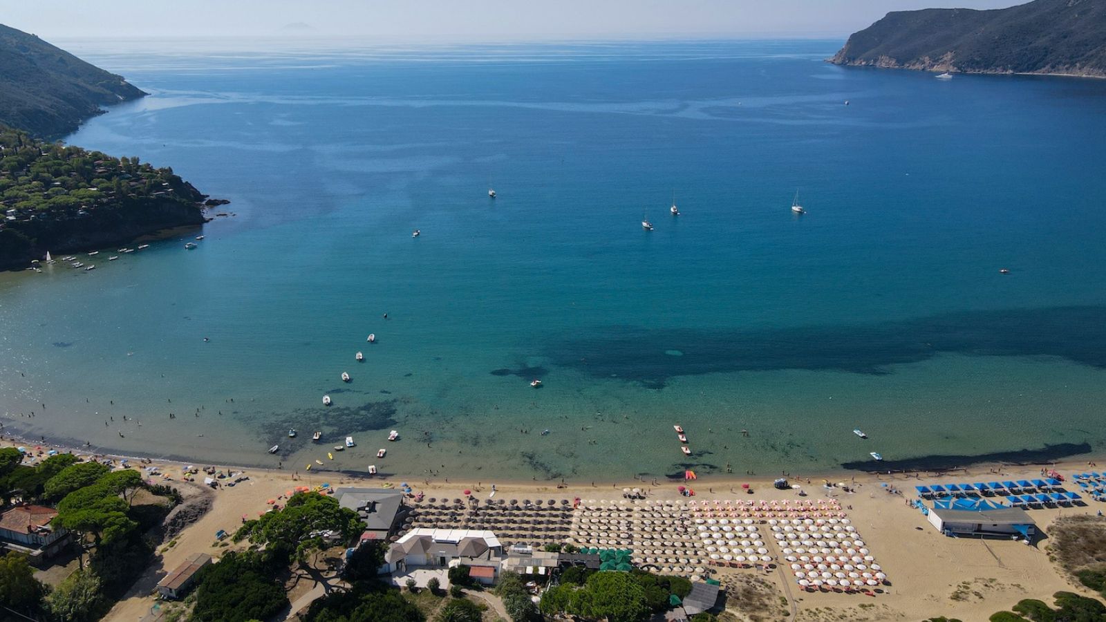 Spiaggia affollata con ombrelloni, barche in mare e colline verdi sullo sfondo, sotto un cielo sereno.