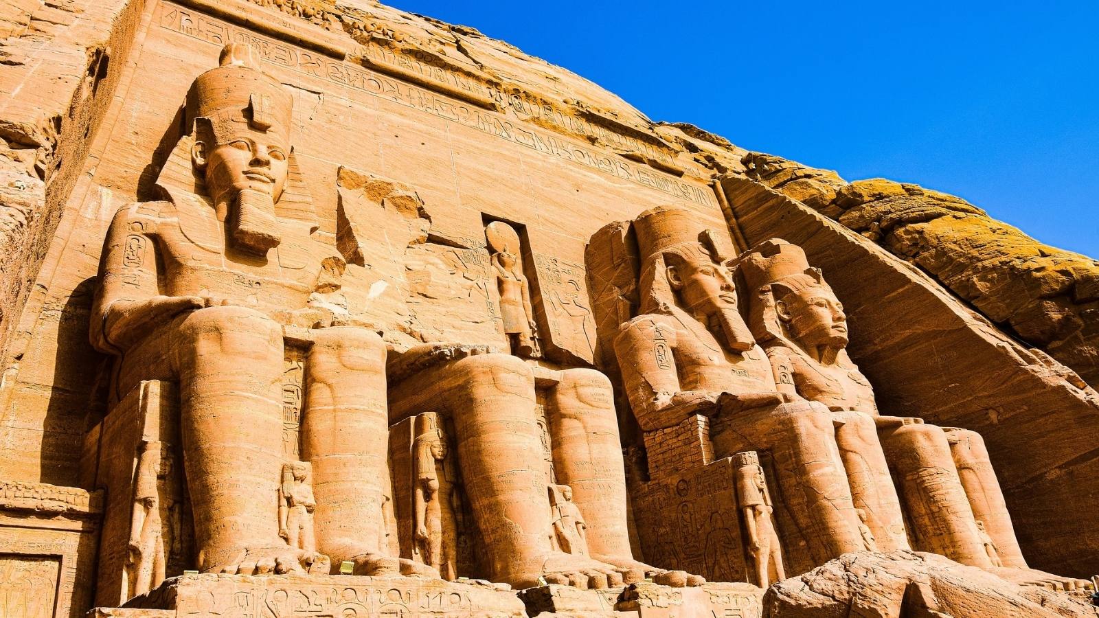 Statue giganti dei faraoni a Abu Simbel, scolpite nella roccia, sotto un cielo blu. Dettagli geroglifici visibili.
