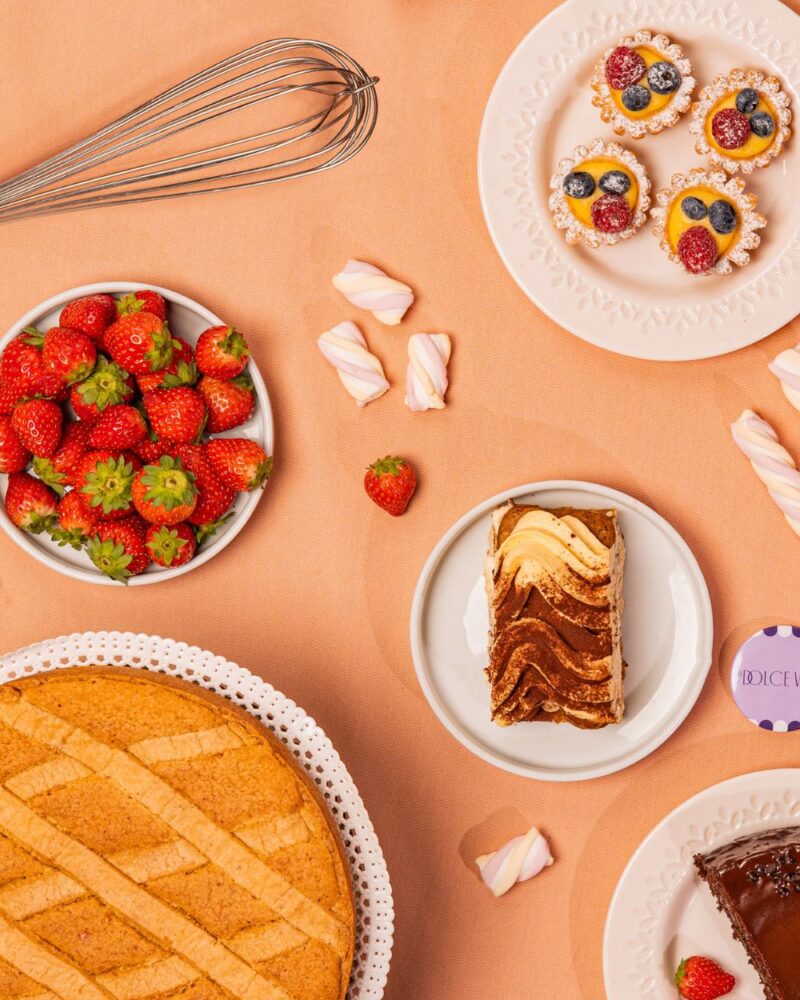 Vari dolci su una tovaglia arancione: torta, dolcetti di frutta, fragole fresche e meringhe. Utensili da cucina in vista.