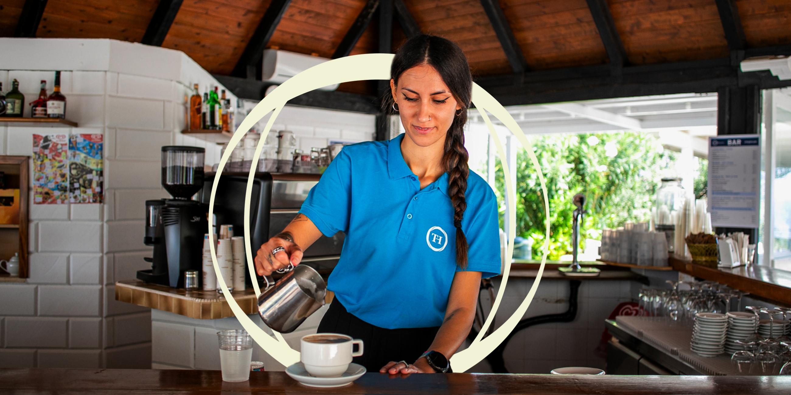 Donna con maglietta blu versa latte in una tazza di caffè al bancone di un bar rustico.