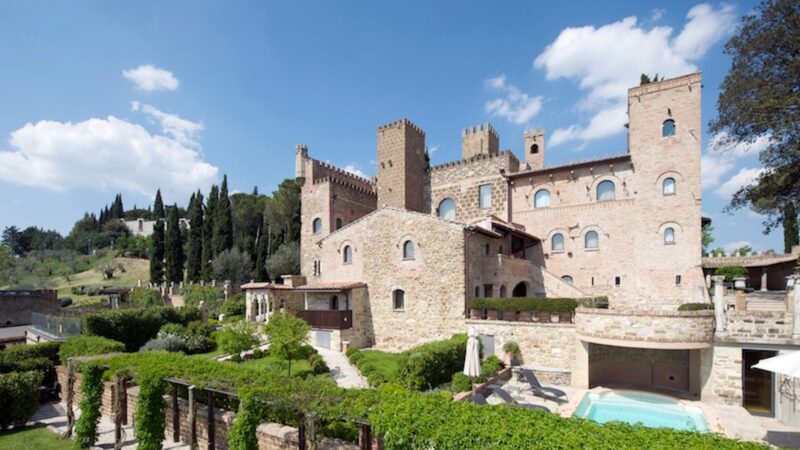 Castello medievale in pietra con giardini curati, piscina e cielo sereno con nuvole.